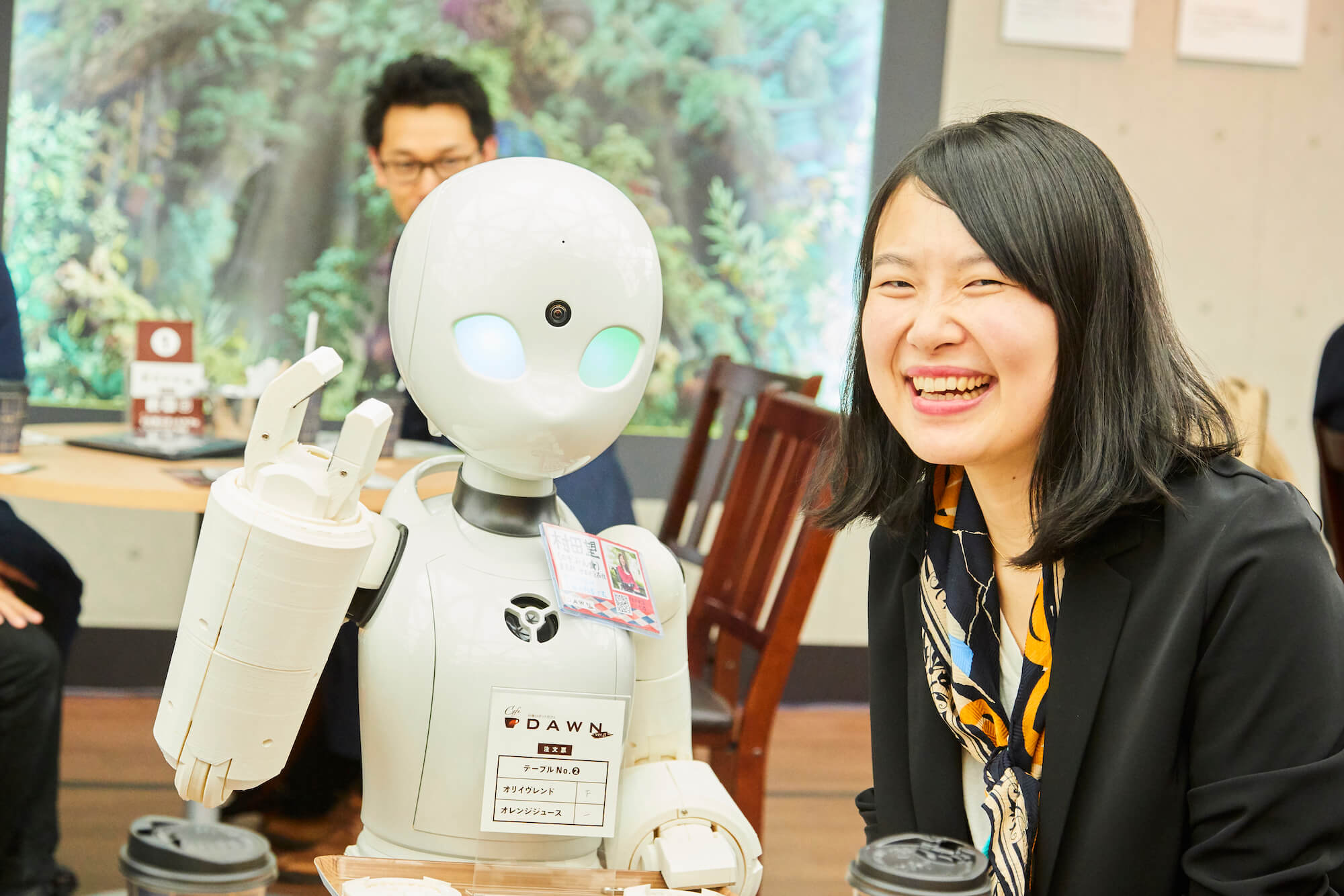 写真：片手をあげる分身ロボットと笑顔の取材記者