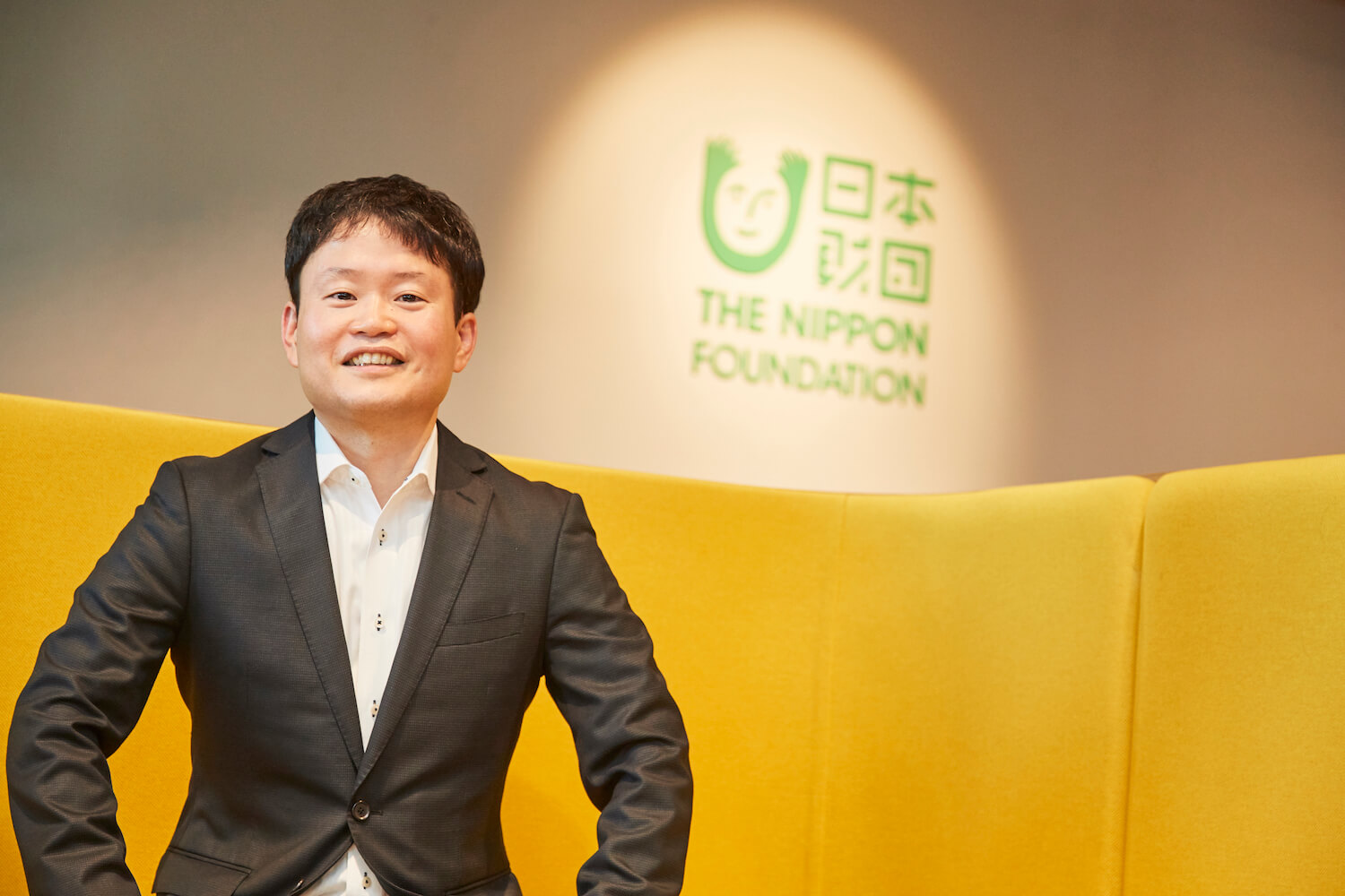 写真：日本財団のロゴを背景に笑顔を浮かべる、日本財団国内事業開発チームのチームリーダー、芳川龍郎さん