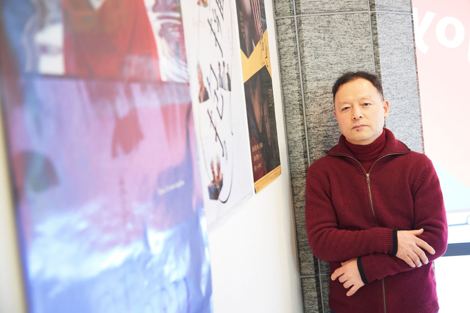 写真:作品のポスターが貼った壁を背景に、腕を組み、真剣な表情を浮かべる坂口香津美さん