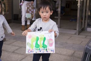 写真：手形と足形を貼り付けた画用紙を持つ男の子