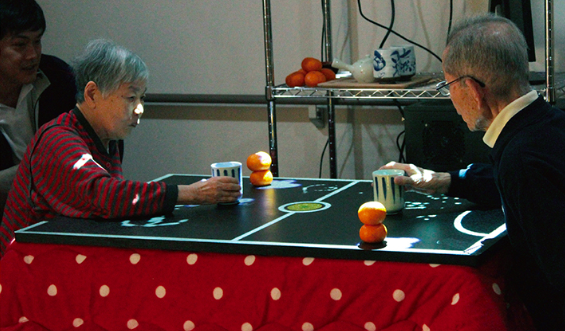 写真：座ったままこたつのテーブルで、デジタル表示されたみかんでホッケーを楽しむ2人の高齢者