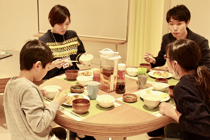写真：料理を囲んで一緒に食事をする2人のスタッフと2人の子ども