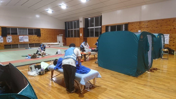 写真：体育館内に置かれた簡易ベッドのうえに横たわった被災者にマッサージをする茨城県鍼灸師会の活動の様子