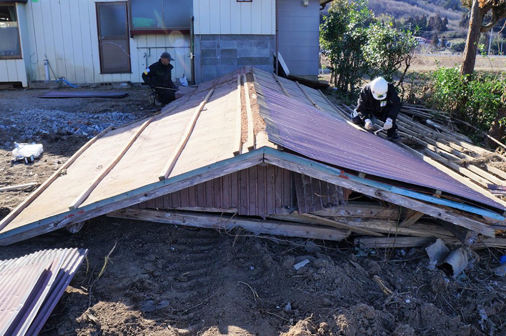 写真：庭にあった小屋の屋根を解体する作業。地面に置かれた大きな屋根の上で男性2人が、しゃがんで釘抜きなどの作業をしていました