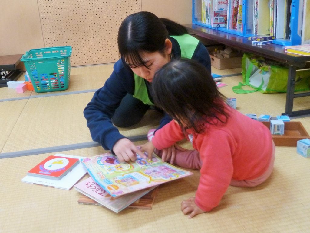 写真：畳の上に座り、被災地の子どもに指で追いながら絵本の読み聞かせをする高校生の様子（12月8日、横沢祥子さん提供）