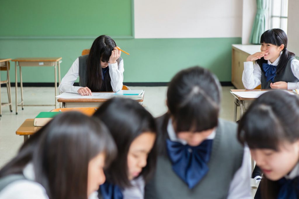 写真：教室の中で1人、机の上で頭を抱える女子高生。その周囲には楽しそうに会話する複数の女子高生の姿が。