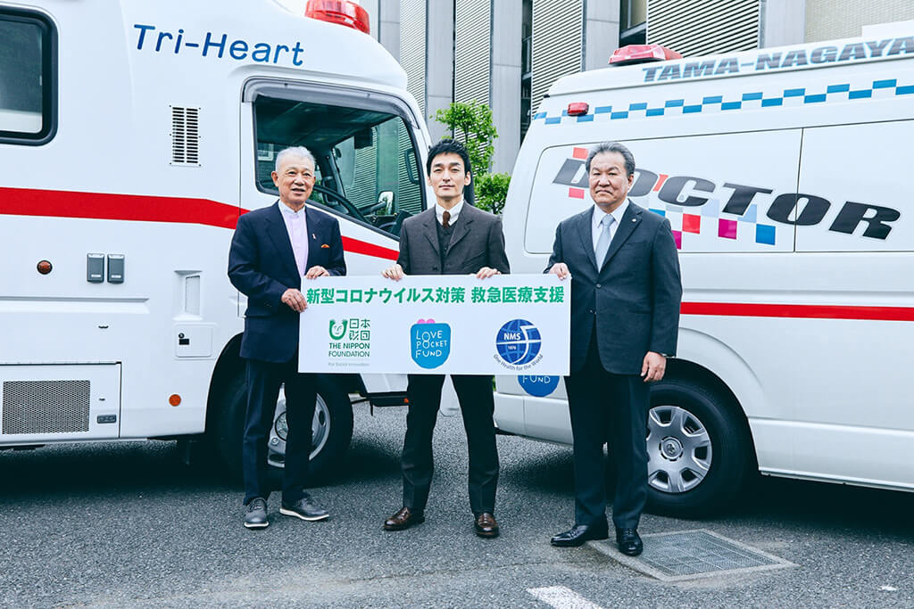 写真：左から笹川、草彅さん、坂本理事長。ドクターカーの前で「新型コロナウイルス対策 緊急医療支援」と書かれたパネルを3人で持つ。