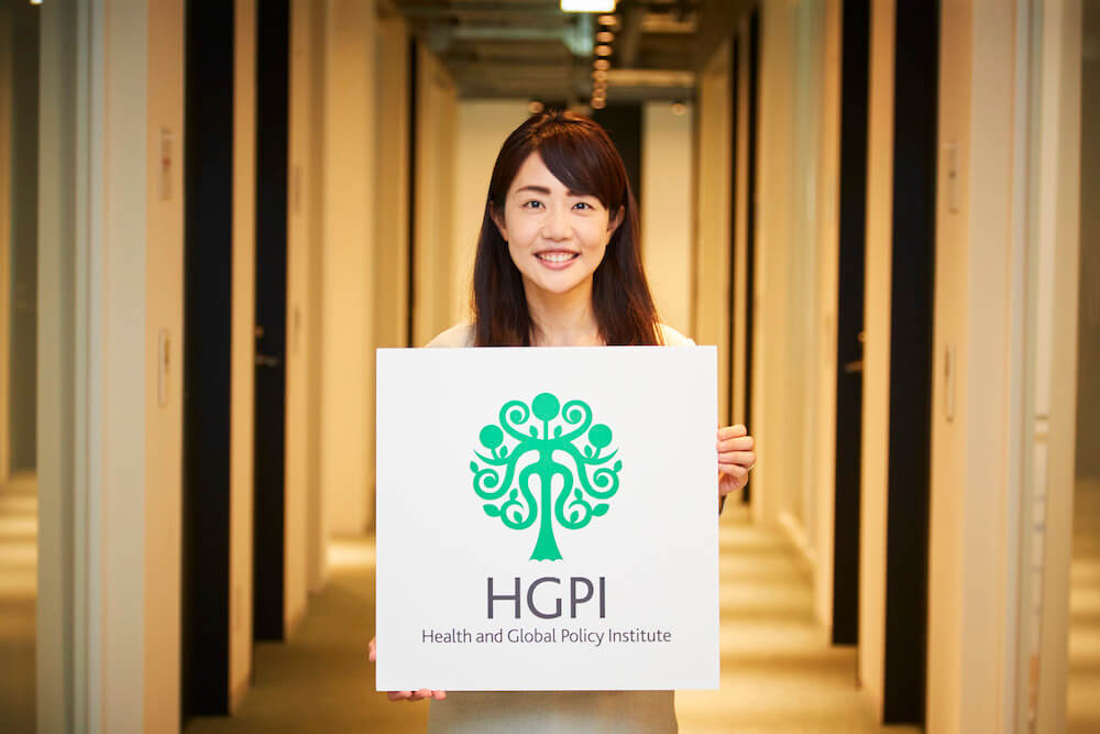 写真：日本医療政策機構のロゴを手に持ち笑顔を向ける今村優子さん