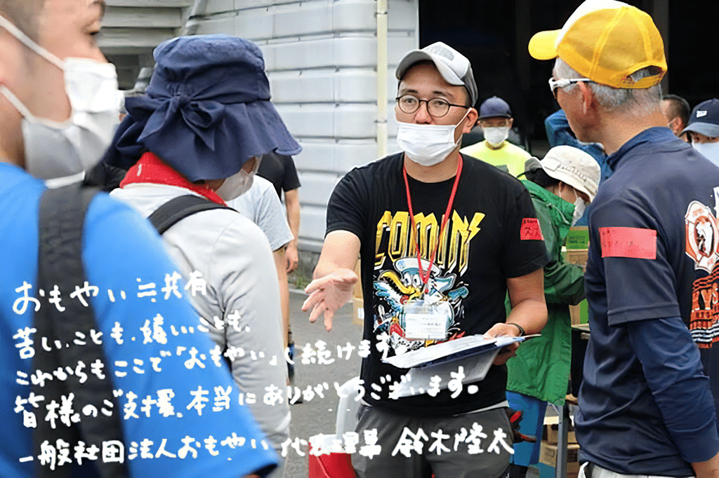 写真：ボランティア参加者たちに話をしている鈴木さん。画面左下に「おもやい≒共有。苦しいことも、嬉しいことも、これからもここで『おもやい』続けます。皆様のご支援、本当にありがとうございます。一般社団法人おもやい 代表理事 鈴木隆太」の文字