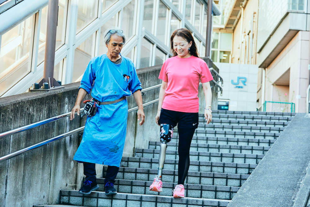 写真：階段を降りる義足の女性（左）と、それを見守る臼井二美男さん