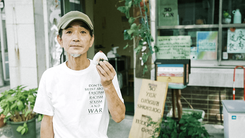 写真：「50円おにぎり食堂」の店先でおにぎりを片手にファインダーに目を向けるヒデさん