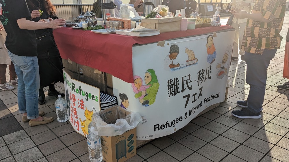 写真：上野駅前に設置された「移民・難民フェス」のパネルが貼られた屋台と、それに集う人々