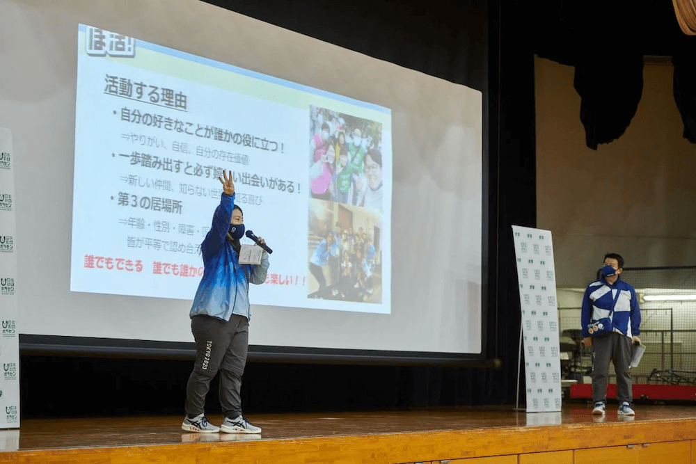 写真：体育館のステージの上でスクルーンに映った資料を説明するボランティアの講師