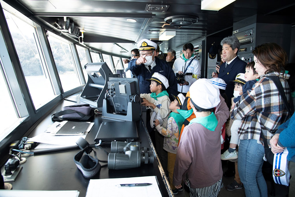 写真：無人運航船の乗船体験で、船長から船の航行について解説を受ける参加者たちの様子。