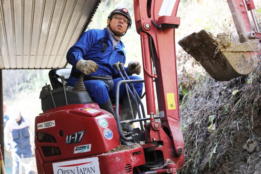 写真:重機を使って家屋の裏に崩れた土砂を撤去する萬代さん(一般社団法人OPEN JAPAN提供)