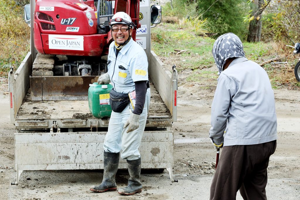 写真:丸森町で重機を背に、作業服姿で住民と話す萬代さん(左)(OPEN JAPAN提供)