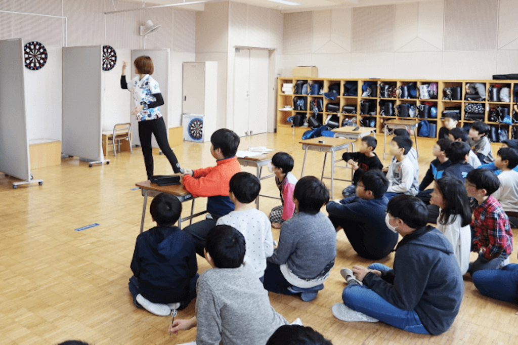 写真:指導スタッフがダーツをする様子を教室で見守るたくさんの子どもたち