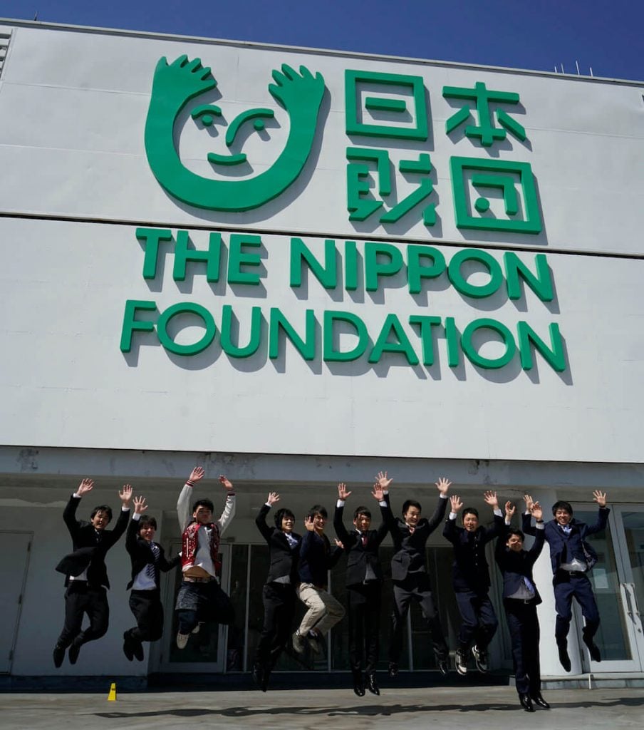 写真:ビルの屋上にある日本財団のロゴマークの前で元気よくジャンプする日本財団夢の奨学金の奨学生たち