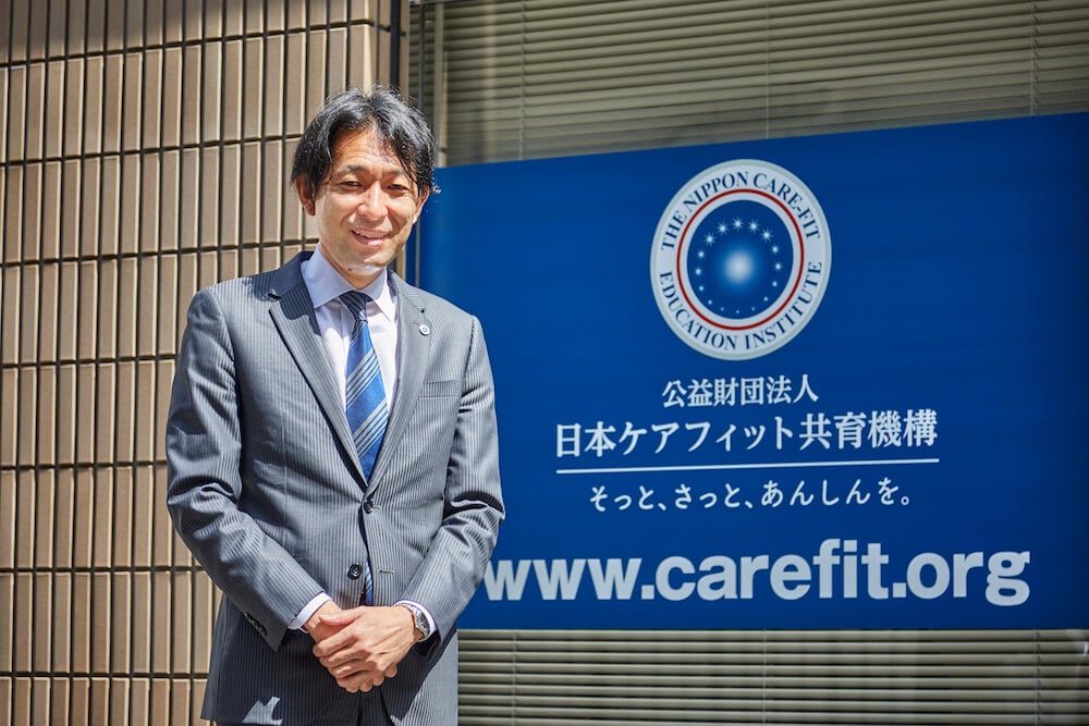 写真:日本ケアフィット共育機構のロゴの前に立つ冨樫さん