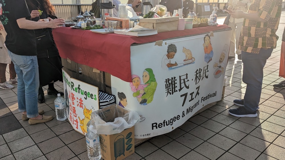 写真:上野駅前に設置された「移民・難民フェス」のパネルが貼られた屋台と、それに集う人々