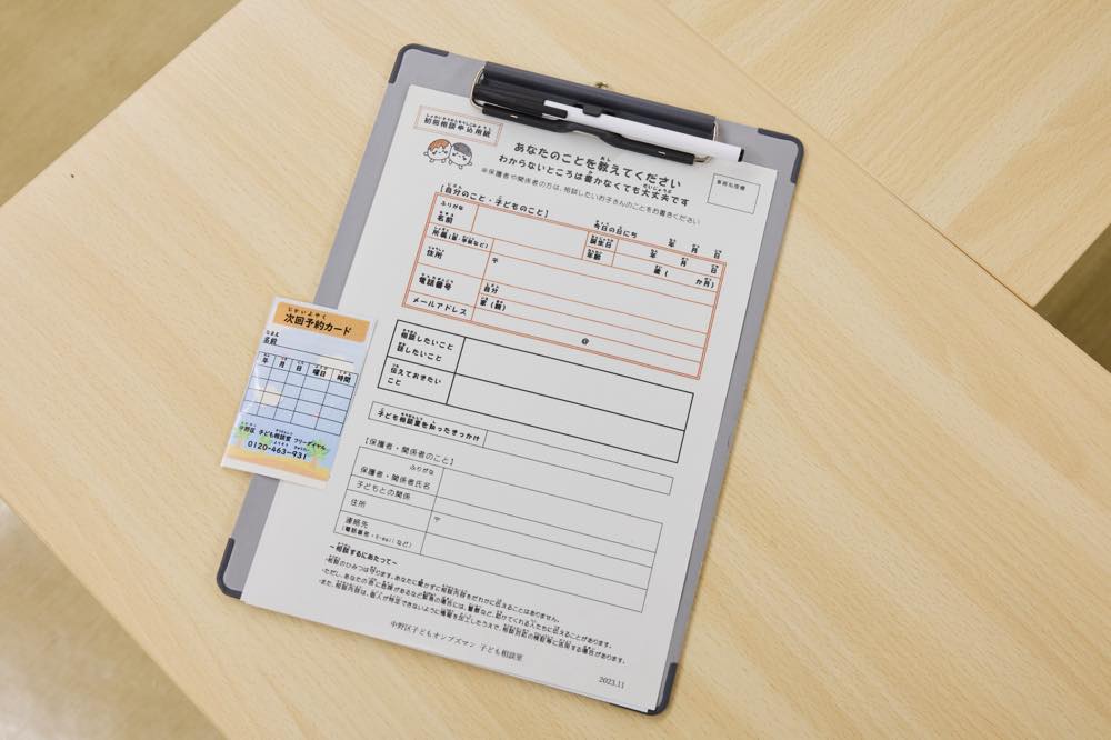 記入用紙「あなたのことを教えてください(わからないところは書かなくても大丈夫です)」の写真。
用紙の上段は基本情報欄で、氏名・ふりがな、年齢、学校・学年、電話番号、メールアドレス、保護者(関係者)の氏名や続柄・連絡先などを書くマスが並ぶ。
その下に自由記述の枠があり、「相談したいこと」「話したいこと」「伝えておきたいこと」などを書く欄が続く。
さらに「子ども相談室を知ったきっかけ」を書く欄、保護者・関係者について(氏名・ふりがな、子どもとの関係、住所、連絡先 など)を書く欄が下部に配置されている。最下段には、記入上の注意や情報の取り扱いに関する説明文が小さく印字されている。
「次回予約カード」には、日付や時間を記入する表と、電話番号(0120-463-931)が印刷されている。
