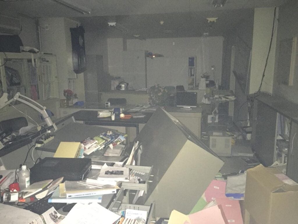 地震で激しく散らかった事務室の内部。大きなキャビネットが倒れ、書類やファイルが床一面に崩れ落ちている