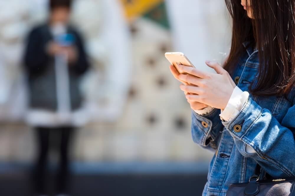 屋外でスマートフォンを両手で操作する、デニムジャケットを着た長髪の女性の横顔