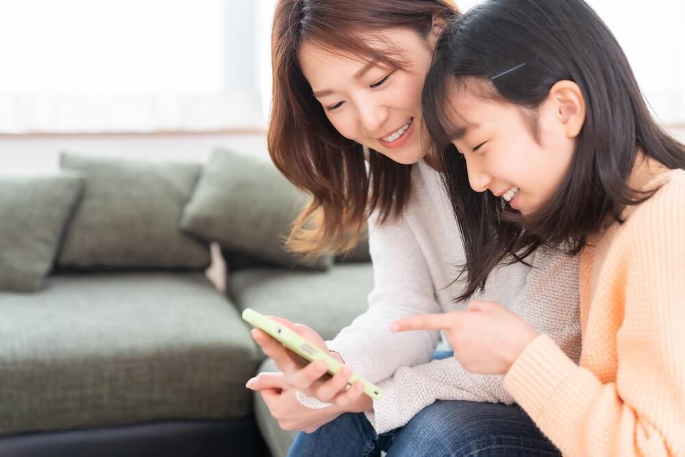 リビングのソファで、笑顔で1台のスマートフォンを一緒に覗き込む母親と小学生くらいの女の子の親子。母親がスマホを持ち、娘が画面を指差しながら楽しそうに話している。