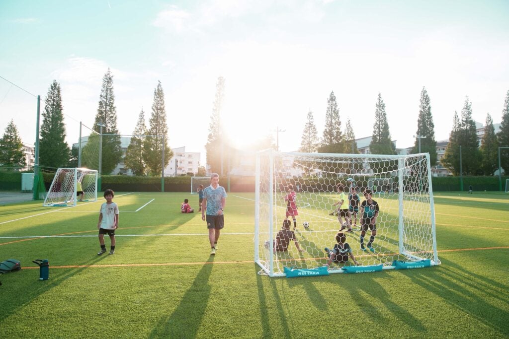 夕暮れ時の柔らかな逆光が差し込む緑の人工芝グラウンドで、サッカーゴールを囲み練習や休憩をする子どもたちと、グラウンドを歩くコーチの姿。