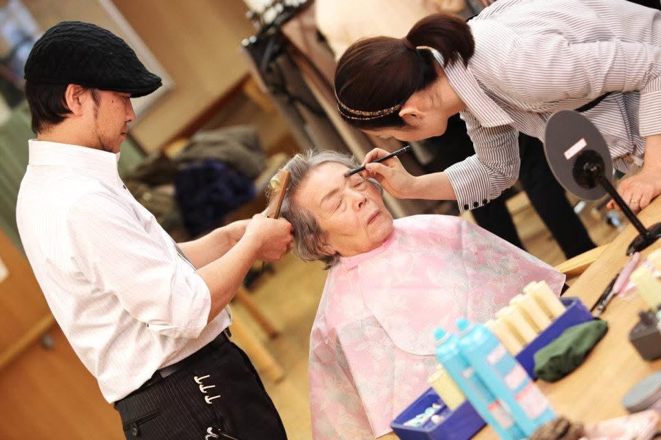 高齢女性が男性理美容師と女性ヘアメイクアップアーティストによる訪問美容サービスを受けている様子