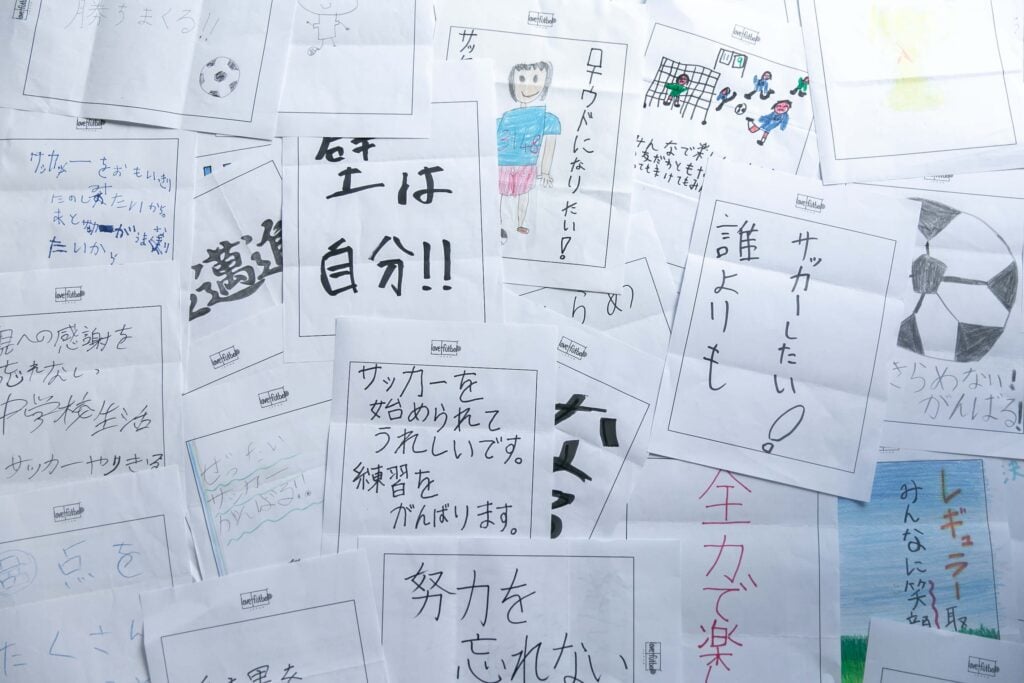 子どもたちが手書きしたメッセージやイラストが描かれた紙が多数敷き詰められている。「壁は自分!!」「誰よりもサッカーしたい!」「サッカーを始められてうれしいです。練習をがんばります。」「努力を忘れない」「ロナウドになりたい!」などの前向きな決意や夢、感謝の言葉が並んでいる。