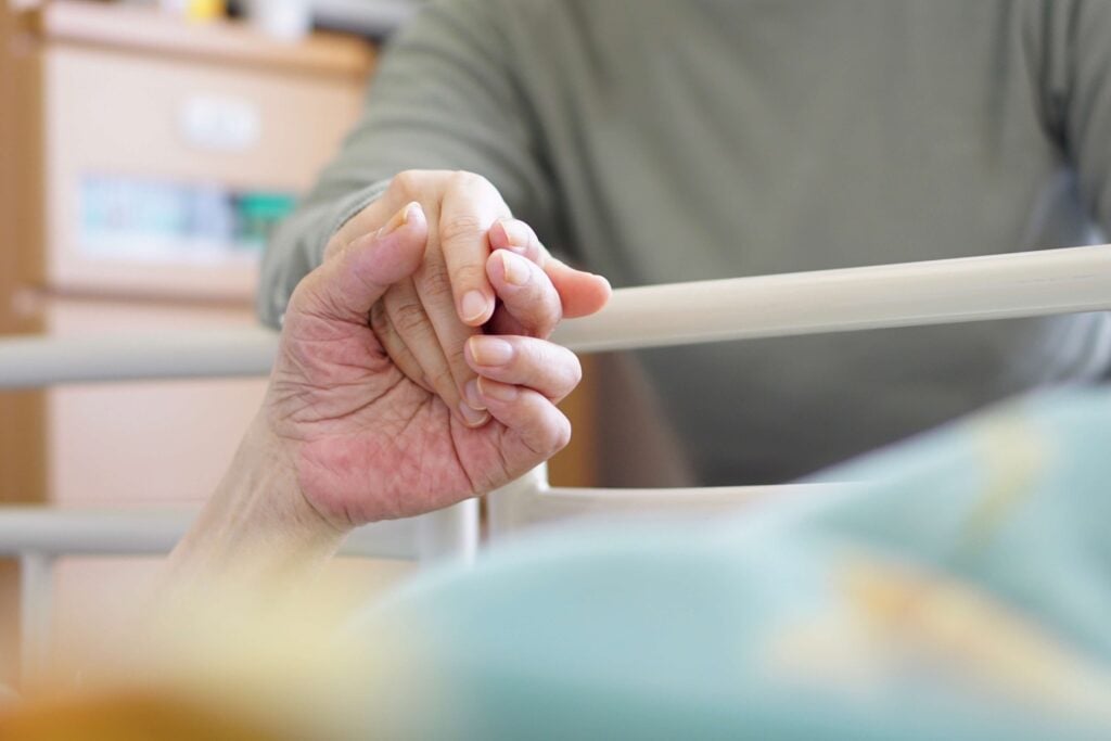 ベッドに横たわる高齢者の手を、寄り添う家族が優しく握りしめる手元の様子
