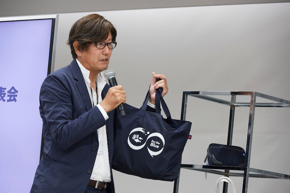 写真：廃棄漁網由来のかばんを手に登壇する兵庫県鞄工業組合・由利理事長